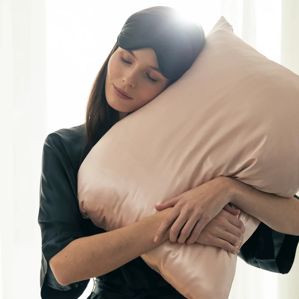 Frau mit Schlafmaske kuschelt ein roséfarbenes Seidenkissen in entspannter Morgenstimmung – Symbol für Schlafkomfort und Wellness.