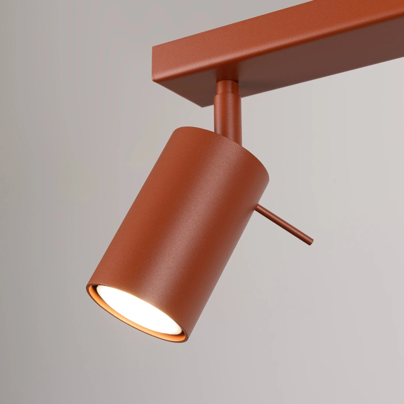 Moderner, rostfarbener zylindrischer Schienenstrahler mit rechteckiger Halterung an hellfarbener Wand.