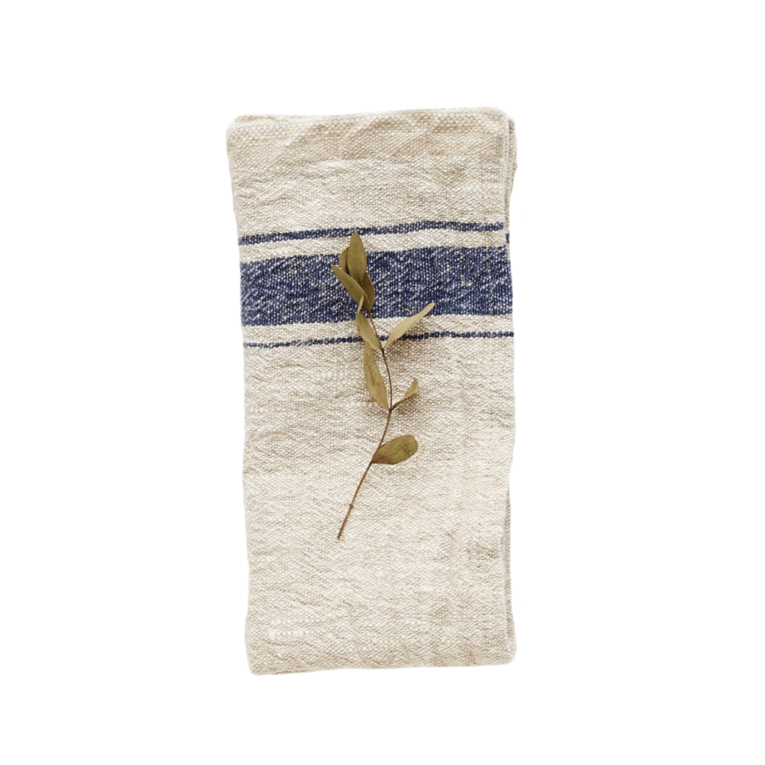 Beiges Leinentuch mit breitem blauen Streifen, rustikaler Struktur und dekorativem Trockenblatt.