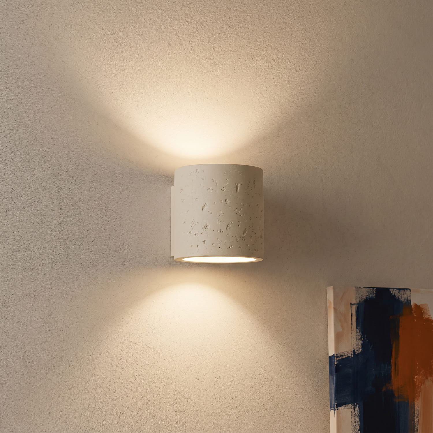 Weiße ORBIS Wandleuchte aus Beton mit warmem Up- und Downlight an strukturierter Wand neben abstraktem Bild